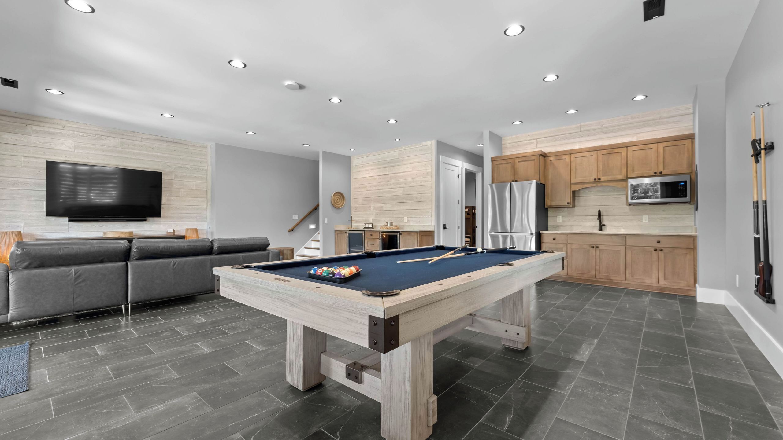 A modern living space with a pool table, grey sofa, wooden cabinets, and a flat-screen TV.