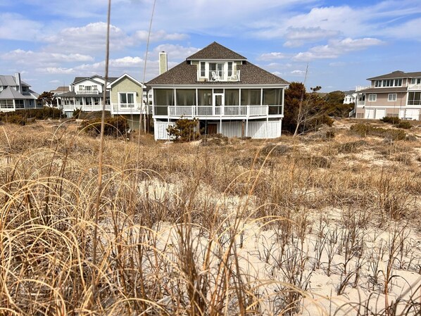 Una casa frente a la playa con un balcón amplio, rodeada de pastizales secos y arena.