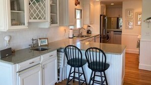 Een keuken met witte kastjes, een centraal eiland met twee zwarte barstoelen, een roestvrijstalen koelkast, en een raam met decoratief traliewerk.