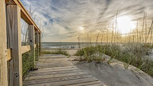 Une promenade en bois menant à une plage, avec un ciel dégagé et le soleil se couchant ou se levant.