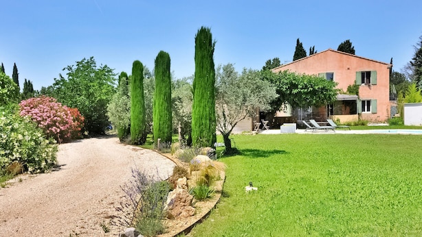 Une maison avec une pelouse bien entretenue, de grands cyprès et un chemin de gravier menant à l’entrée.