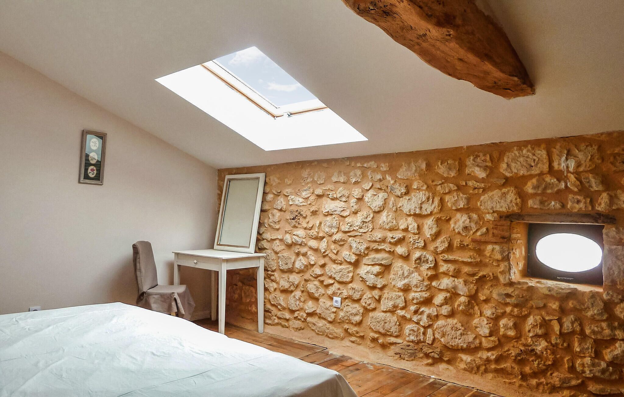 Un dormitorio con pared de piedra, tragaluz, cama, silla y ventana pequeña.