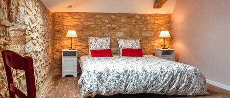 A bedroom with a stone wall, a wooden ceiling, a bed with floral bedding, two bedside lamps, and a red chair.