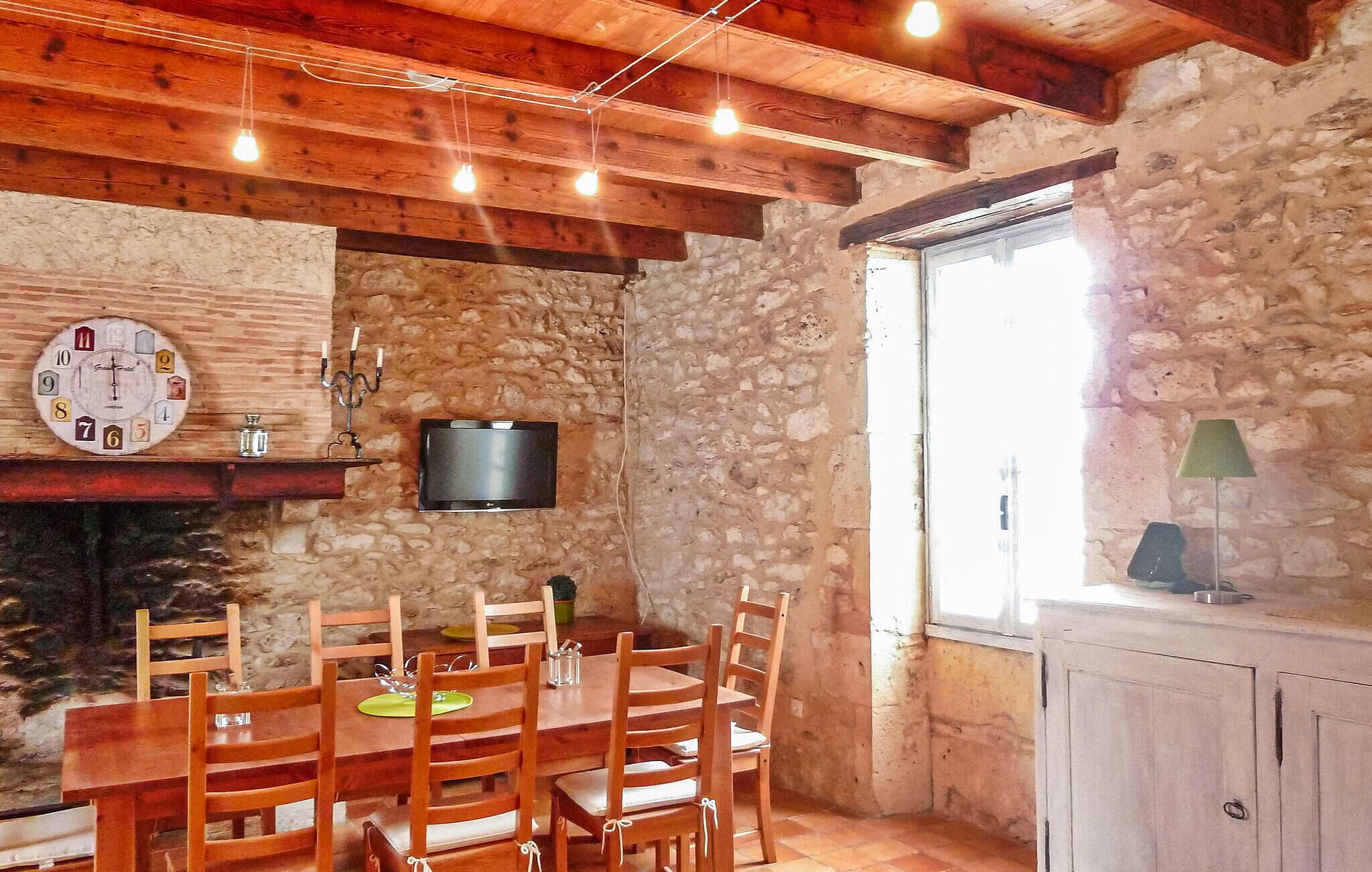 A rustic dining area with a stone wall, wooden ceiling beams, a clock, a flat-screen TV, and a window with a view.