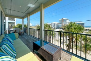 A balcony with a wicker table and a sofa, overlooking a beach and buildings. - Sandy Cheeks | Outdoor Pool | Waterfront | My Beach Getaways (Gulf Shores)