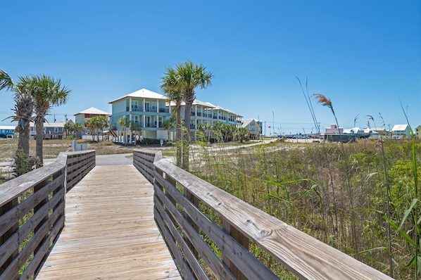 A wooden boardwalk leading to a beachfront property with multiple buildings, palm trees, and a clear blue sky. - Sandy Cheeks | Outdoor Pool | Waterfront | My Beach Getaways (Gulf Shores)