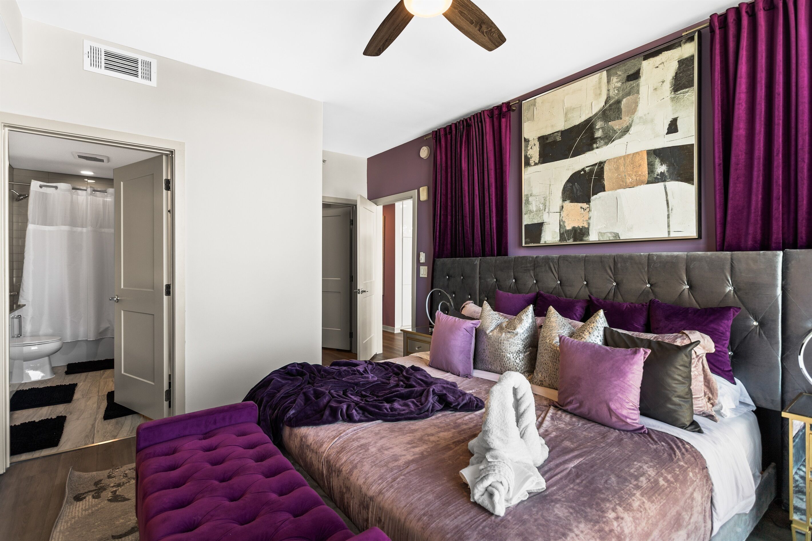 A bedroom with a tufted headboard, a large bed with multiple pillows, a bathroom with a shower curtain, and a ceiling fan.