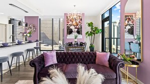 A modern living room with a purple sofa, a dining area with a chandelier, and a large mirror reflecting the room. - The Velvet Room | 17th Floor Luxury Condo | Skyline Views & Balcony (Atlanta)