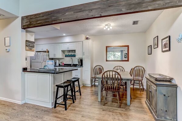 A kitchen with white cabinets, a central island with stools, and a dining area with wooden chairs and a table.