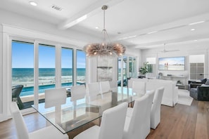 A modern interior with a glass table, white chairs, a chandelier, and a large window overlooking the ocean.
