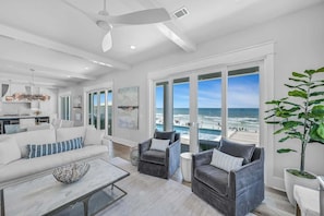 A spacious living area with a large window overlooking the beach, a ceiling fan, and a modern kitchen in the background.