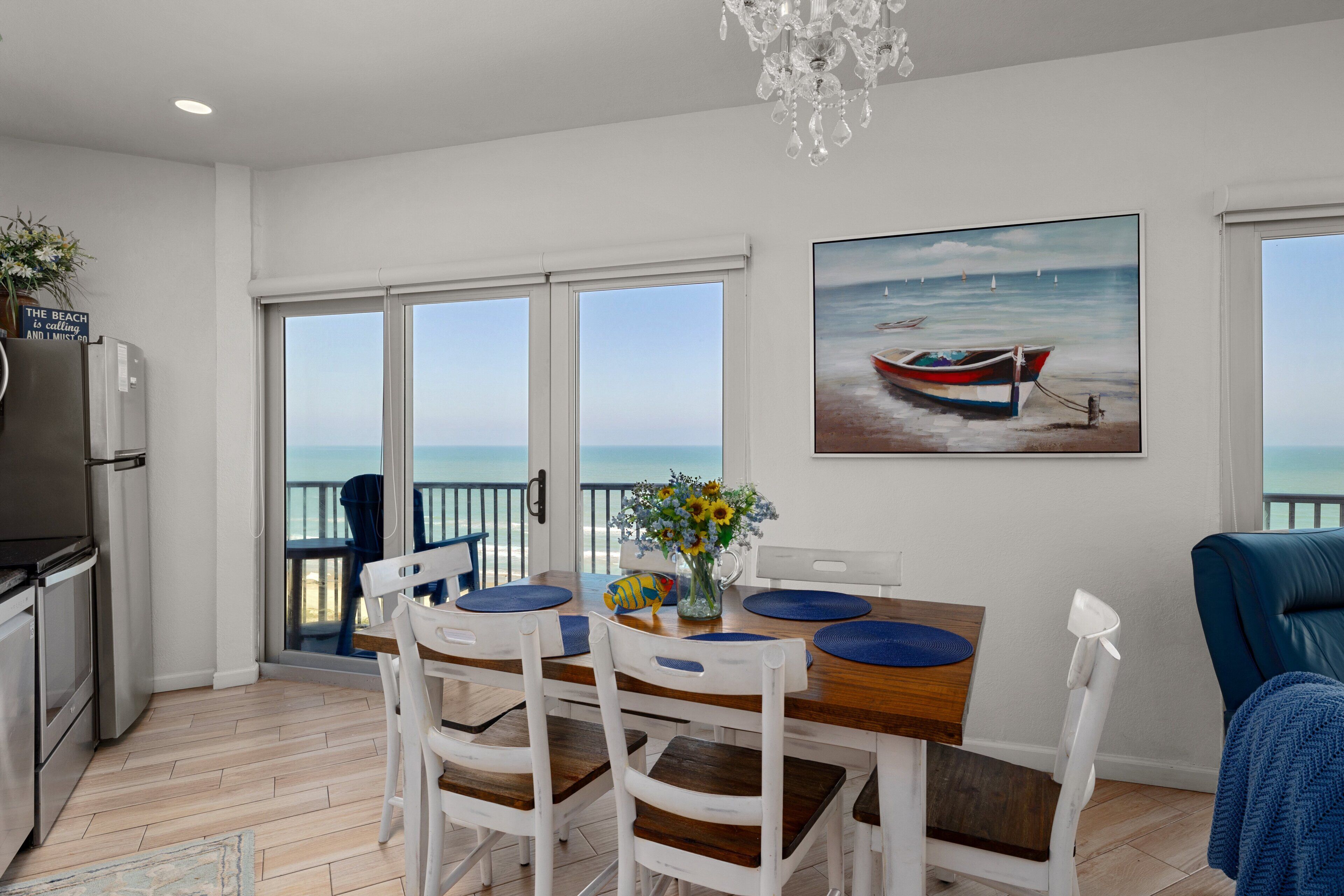 A dining area with a view of the sea, a painting of a boat, and a refrigerator.
