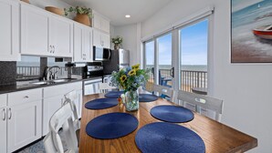 Una cocina moderna con armarios blancos, una mesa de comedor de madera con manteles individuales azules, un jarrón con flores y un ventanal con vistas al océano.