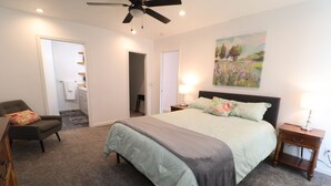 Une chambre à coucher comprenant un lit, une chaise, une table de chevet, un ventilateur de plafond et un tableau accroché au mur.