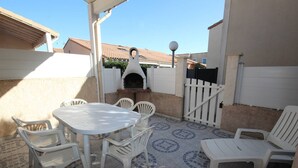 Une terrasse extérieure avec une table ronde et des chaises en plastique blanches, un four à pizza et un portail blanc.