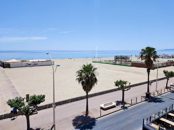A beachside view with a clear sky, a sandy beach, palm trees, and a road with a sidewalk. - Lovely flat sea view, wifi, car park, air conditioning. (CANET PLAGE)