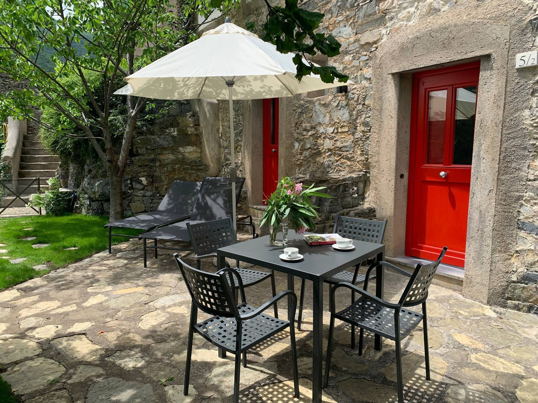 A stone building with a red door, a patio with a table and chairs, and a white umbrella.