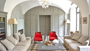 Una sala de estar con dos sofás beige, una mesa de centro y una araña. Hay dos sillones rojos y un amplio arco de entrada.