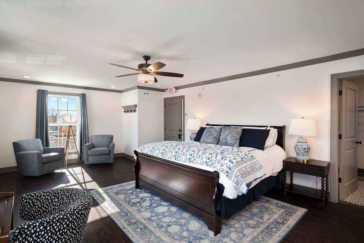 A bedroom with a bed, two armchairs, a ceiling fan, a window with curtains, and a nightstand with a lamp.
