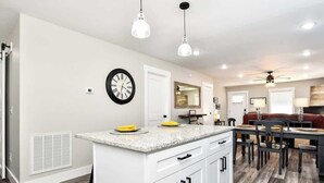 A modern kitchen with white cabinetry, a granite countertop, and a built-in stove. There is a dining area with a red upholstered bench and a chandelier.
