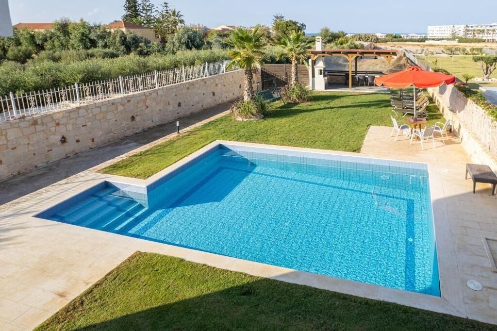 A rectangular swimming pool with a tiled deck, surrounded by a stone wall and a grassy area.