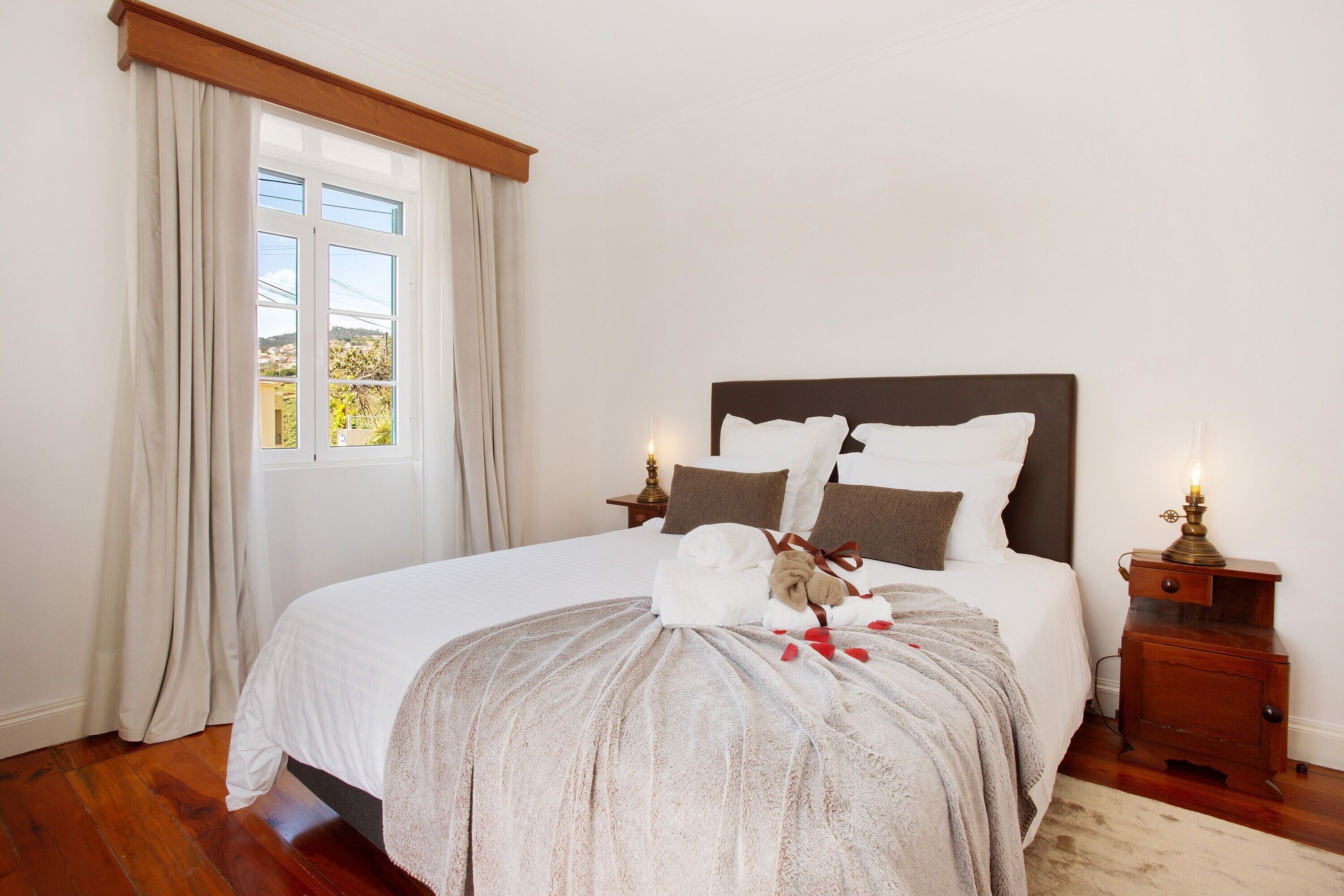 Una cama bien hecha con cabecera, una mesita de noche con lámpara y una ventana con cortinas.