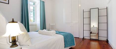 A bedroom with a bed, a bedside table with a lamp, a window with curtains, a wardrobe, and a mirror.