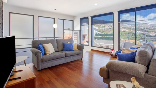 Una sala de estar moderna con un sofá grande, un televisor y vistas a un pueblo a través de amplias ventanas.