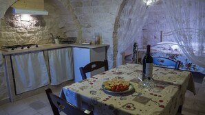 A stone-walled room with a kitchen area, a dining table with a floral tablecloth, and a bottle of wine. - Apartment 'Trullo Fico' with Shared Pool, Shared Terrace and Wi-Fi (Martina Franca)