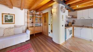 A cozy room with a wooden ceiling, a sofa, a small kitchen, and a wooden shelf with books and decorative items. - Apartment 'Villa Cleila Blu D' with Mountain View, Shared Garden and Balcony (San Vito di Cadore)