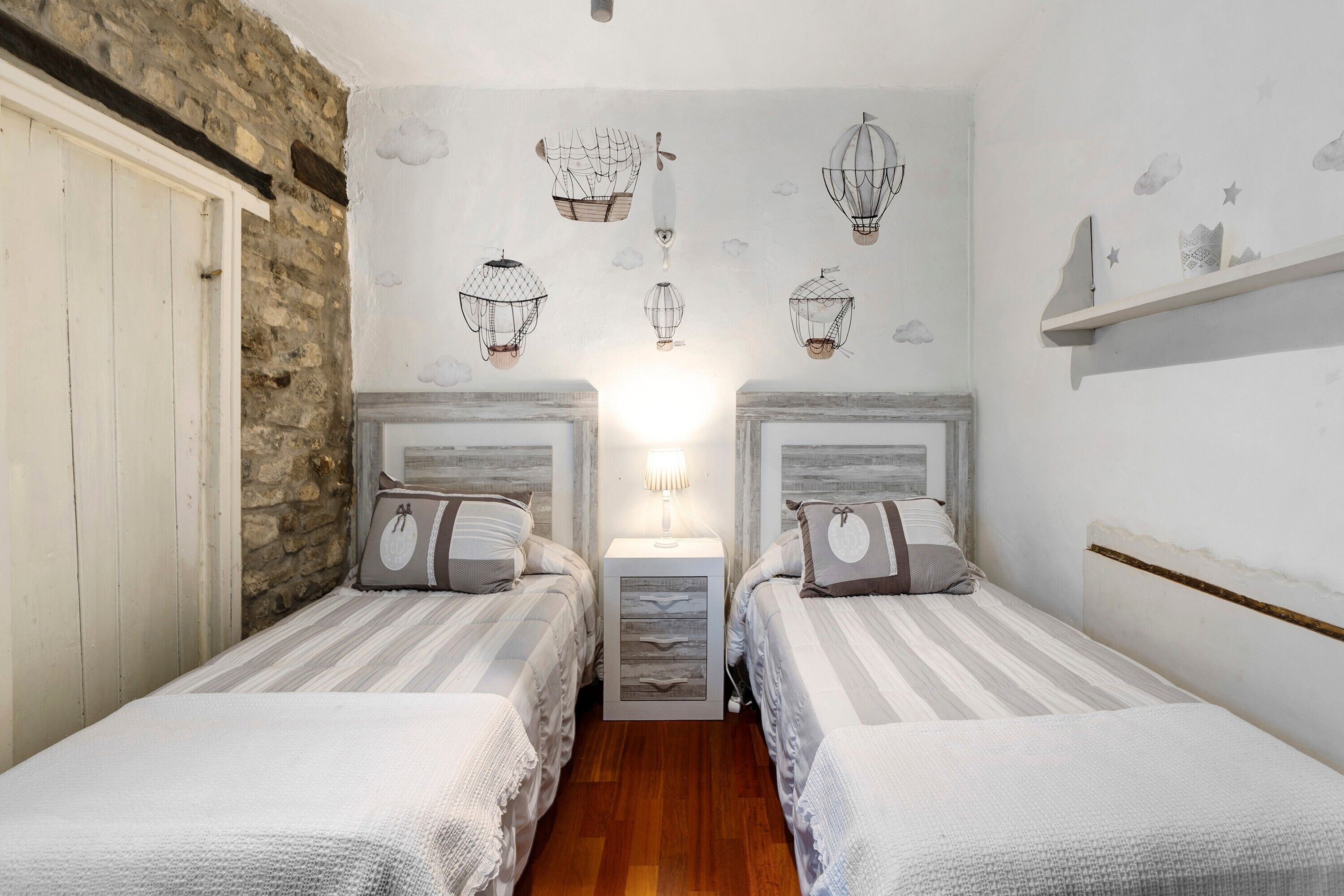 A bedroom with two beds, a nightstand, a wall-mounted lamp, and a shelf with decorative items.