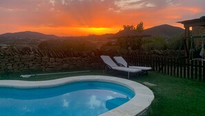 Una piscina con sillones de descanso, un muro de piedra y una vista del atardecer sobre un paisaje montañoso.
