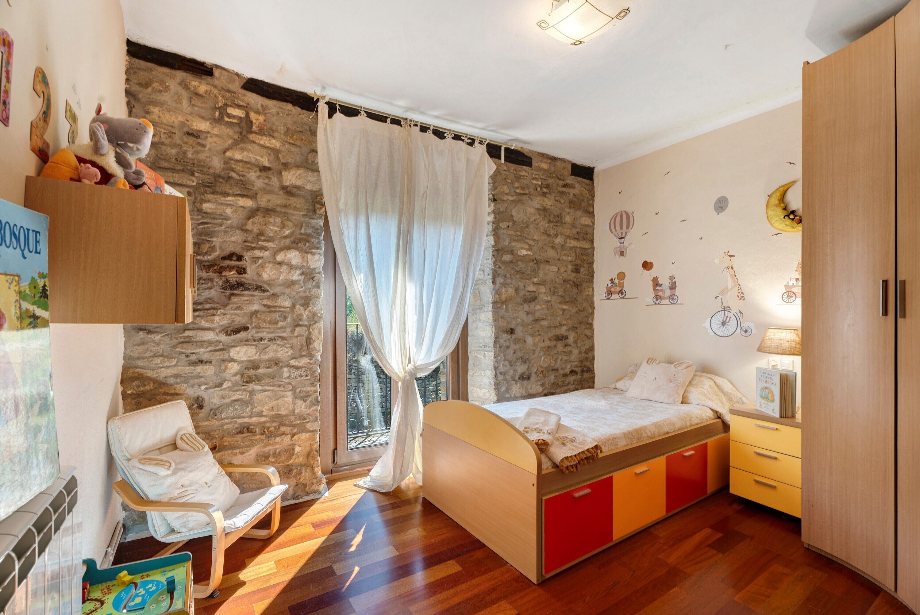 A bedroom with a stone wall, a bed with red and yellow drawers, a white chair, and a window with curtains.