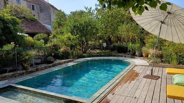 A swimming pool with a wooden deck, surrounded by greenery and a building. - Le Tonnelier – Charming House with Pool in the Heart of Quercy (Limogne-en-Quercy)