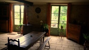 A room with a wooden bench, a table, and a dresser. There are windows with curtains, and potted plants are visible. - Le Tonnelier – Charming House with Pool in the Heart of Quercy (Limogne-en-Quercy)