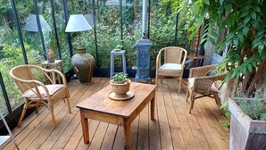 A wooden deck with wicker chairs and a table, surrounded by greenery and a glass enclosure. - Le Tonnelier – Charming House with Pool in the Heart of Quercy (Limogne-en-Quercy)