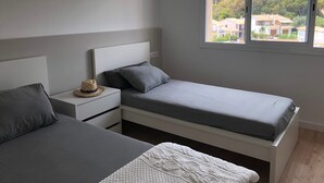 A bedroom with two beds, a nightstand, a hat, and a window with a view of buildings and trees. - Apartment 'Apartament Creu De Sant Pol' with Sea View and Wi-Fi (Sant Feliu de Guíxols)