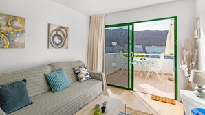 Una sala de estar con sofá, una mesa de centro y un gran ventanal corredizo que da a un balcón exterior con mesa y sillas.