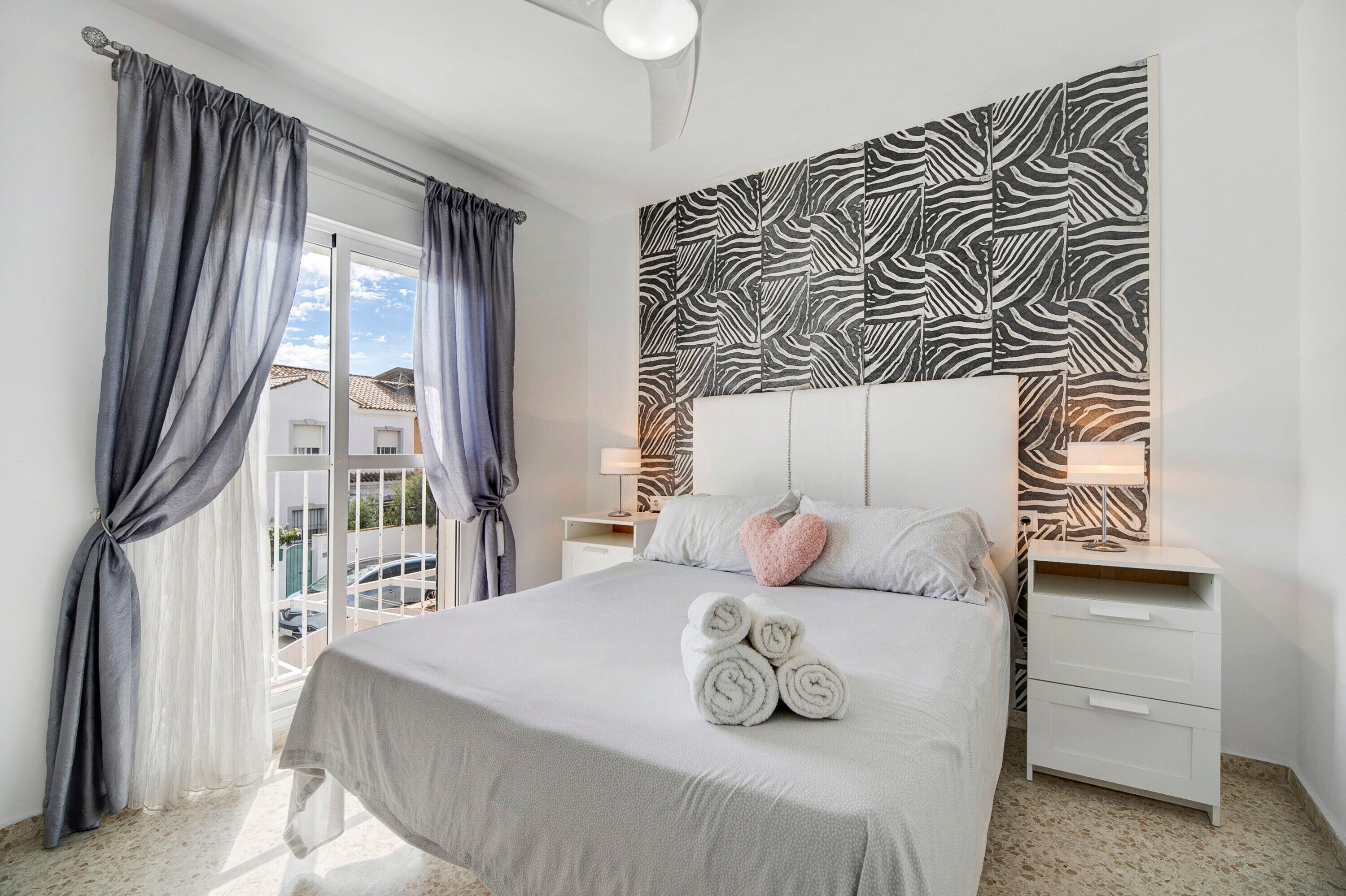 Habitación de hotel con cama, cortinas, un papel tapiz a rayas de cebra, una mesita de noche y una ventana con vistas.