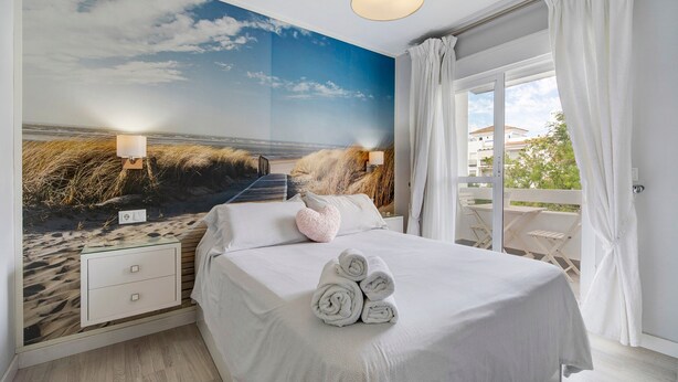 Een slaapkamer met een strandmuur, een bed met opgerolde handdoeken, een nachtkastje en een raam met gordijnen.