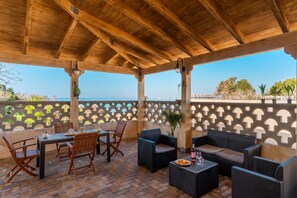 Eine Terrasse im Freien mit Holzmöbeln, einem Tisch mit Flasche und Gläsern und Blick auf das Meer.