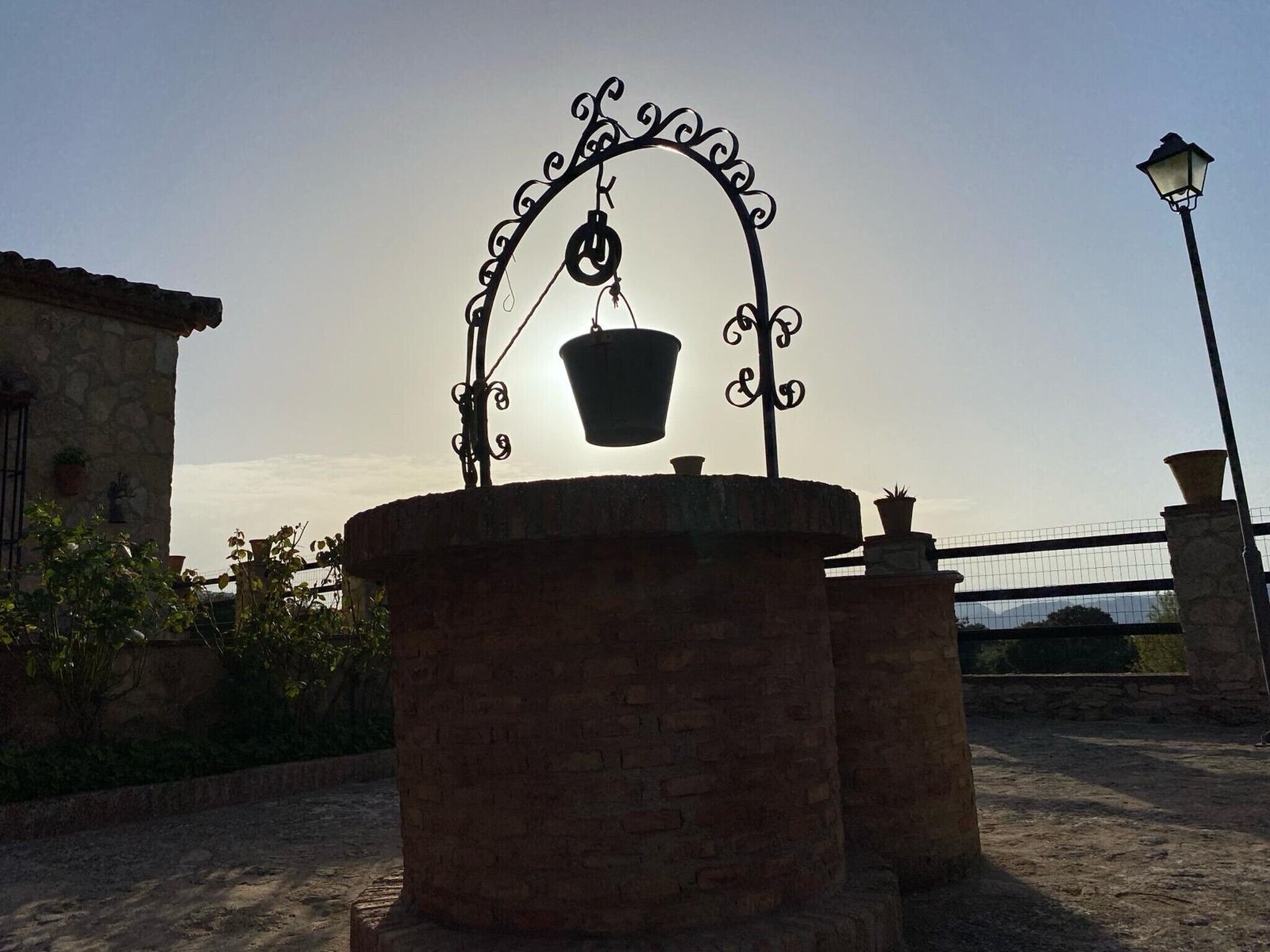 A stone fountain with a metal arch and a hanging bucket, surrounded by brick walls and potted plants.