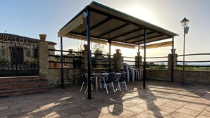 Une terrasse extérieure avec un sol en pierre, des tables et des chaises en métal, un espace salon couvert et un mur en pierre agrémenté d’un portail en métal.