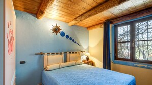 Ein Schlafzimmer mit Holzbalkendecke, einem Bett mit blauer Bettdecke, einem Nachttisch mit Lampe und einem Fenster mit Blick auf Bäume.