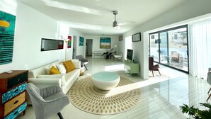 Uma sala de estar moderna com um sofá branco, uma poltrona cinza, uma mesa de centro redonda e uma janela grande com vista para o exterior.