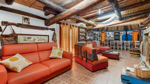 A rustic interior with wooden beams, a sofa, a red armchair, and a dining area with chairs and a table.