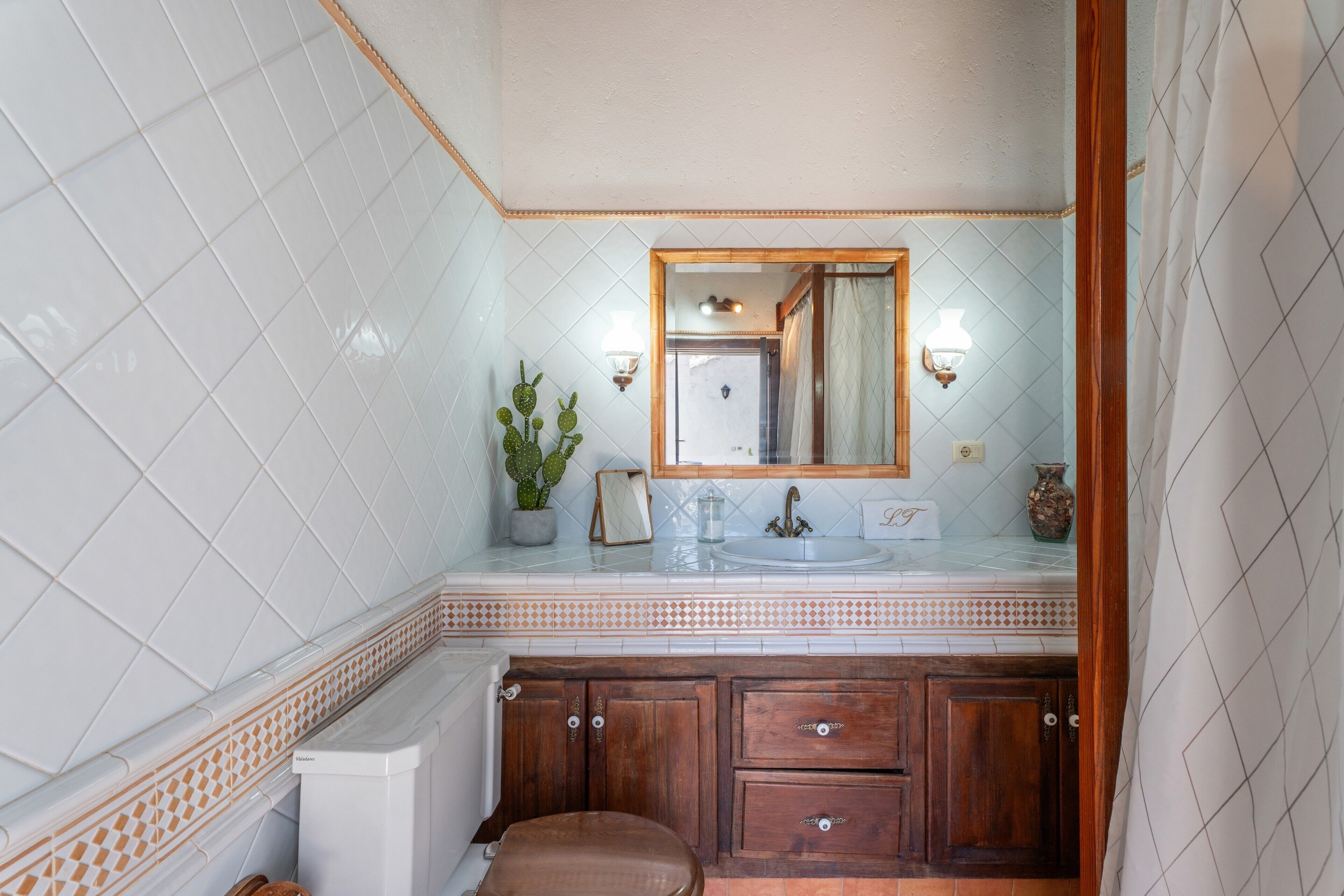 A bathroom with white tiled walls, a wooden vanity, a sink with a mirror, and a potted cactus.