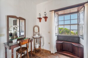 A room with a wooden bench, a table with a chair, a mirror, and a window with a view of greenery. - Casa Rural La Tunera. Historic Canarian Home (1797), Sea & Mountain Views, Pool, Wi-Fi (Villa de Arico)