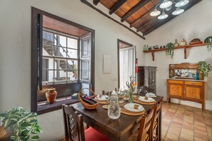 A dining area with a wooden table set for four, a window with a view of a staircase, and a kitchen area with shelves above the counter. - Casa Rural La Tunera. Historic Canarian Home (1797), Sea & Mountain Views, Pool, Wi-Fi (Villa de Arico)