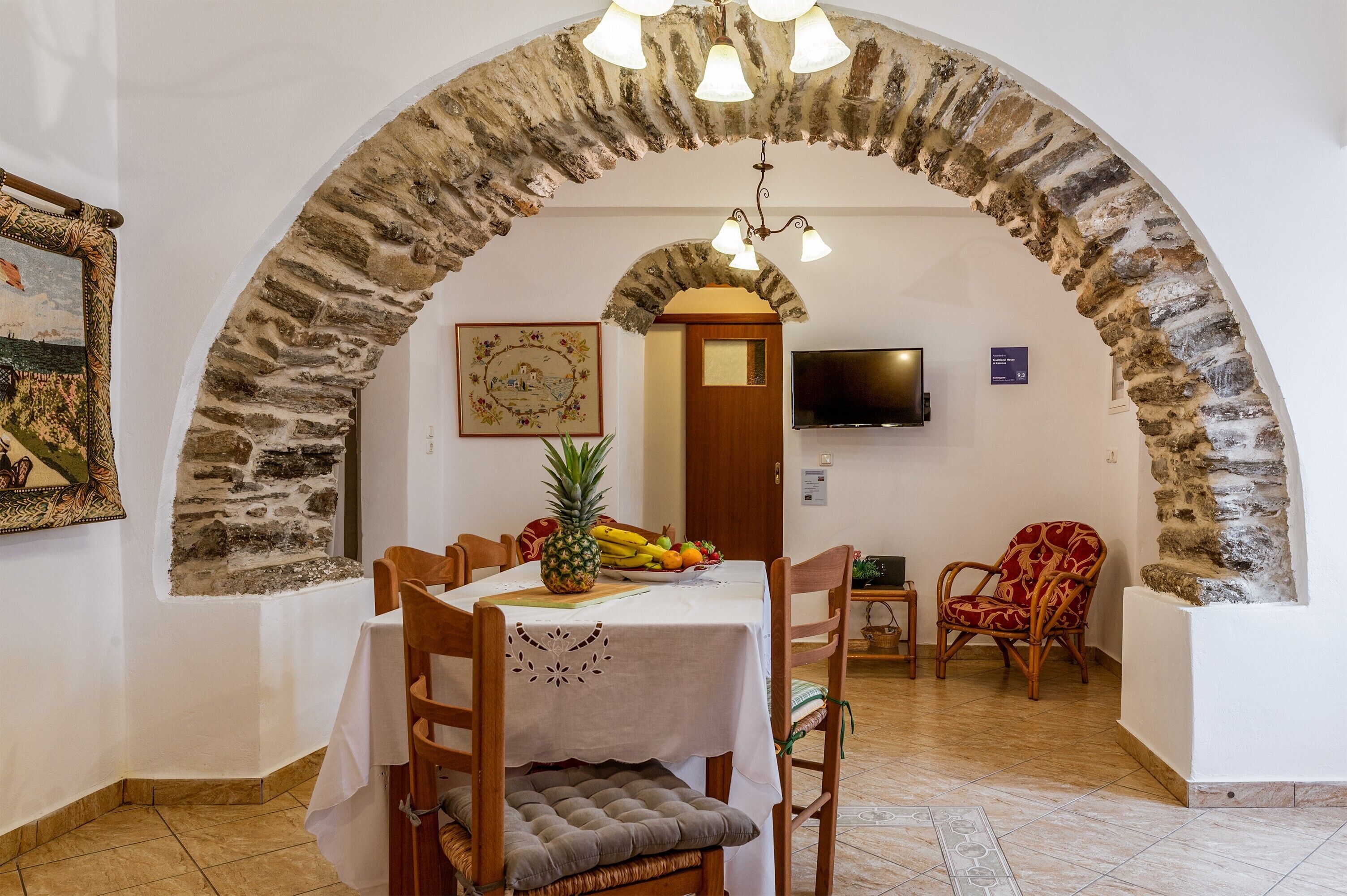 A dining area with a stone arch, a table with a pineapple and fruit, wooden chairs, and a television on the wall.